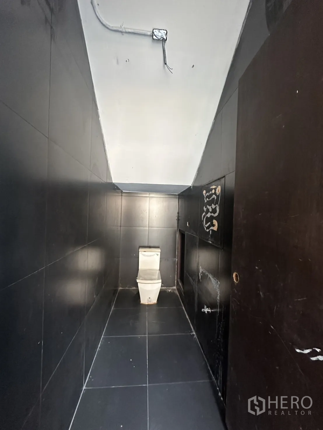 Home Office for rent in Pathum Wan, Bangkok - Narrow black-tiled restroom with a modern toilet and exposed ceiling conduit.