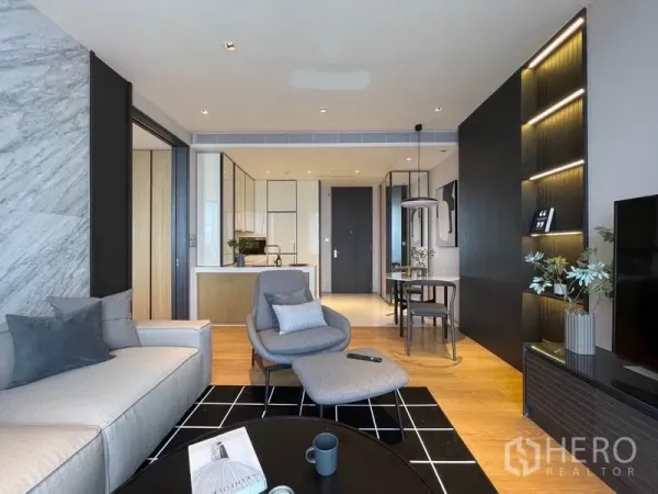 Condominium for rent in Khlong Toei, Bangkok - Living area with lounge chair, black rug, built-in shelving and a sleek white kitchen beyond.