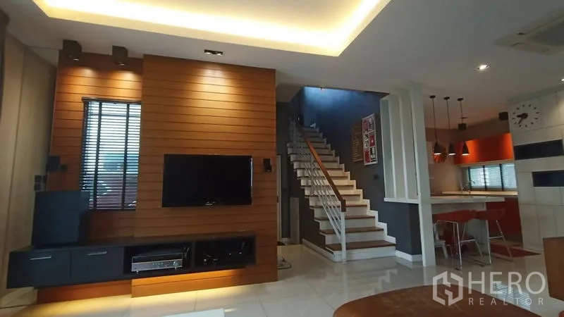 Detached House for sale in Prawet, Bangkok - Open-plan living area with wood accent TV wall, staircase, and kitchen with pendant lights.