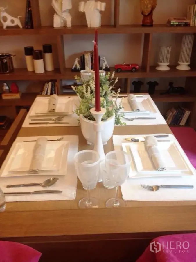 Condominium for rent in Watthana, Bangkok - Dining table set for four with white tableware and a red candle centerpiece, backed by wooden shelves with decor.