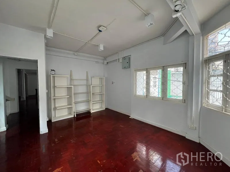 Home Office for rent in Watthana, Bangkok - Bright room with white walls, open shelving, windows with security grilles, and polished wooden floor.