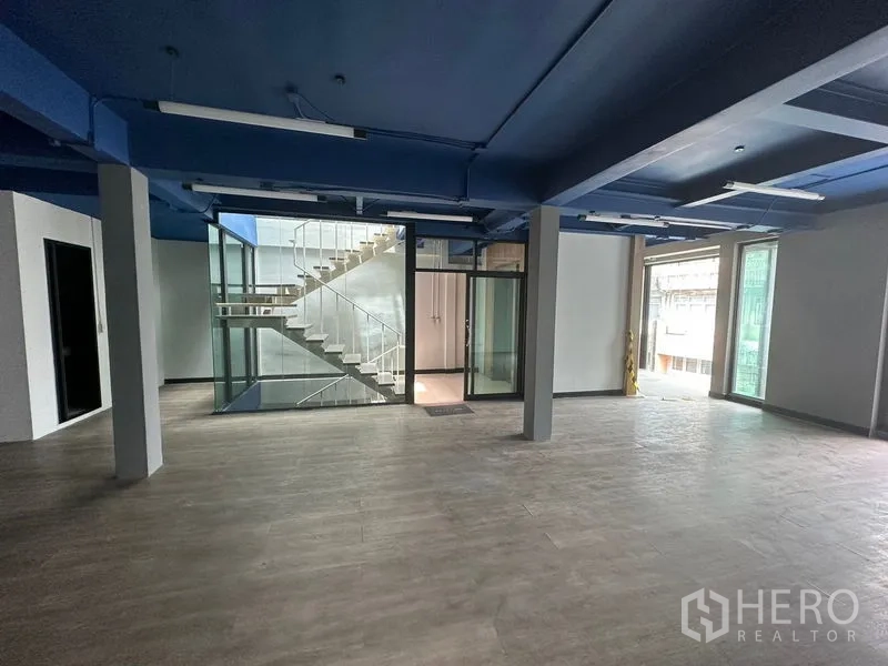 Office Space for rent in Watthana, Bangkok - Open-plan office with blue ceiling, wood-look floors and a glass-walled staircase core.
