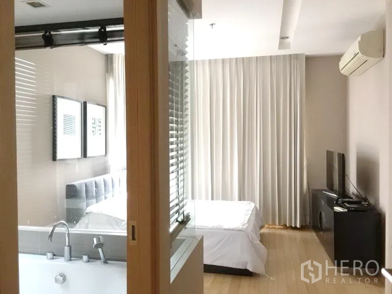 Condominium for rent in Khlong Toei, Bangkok - Master bedroom with glass-partition bathroom and bathtub, bed, TV console and wall-mounted air conditioner.