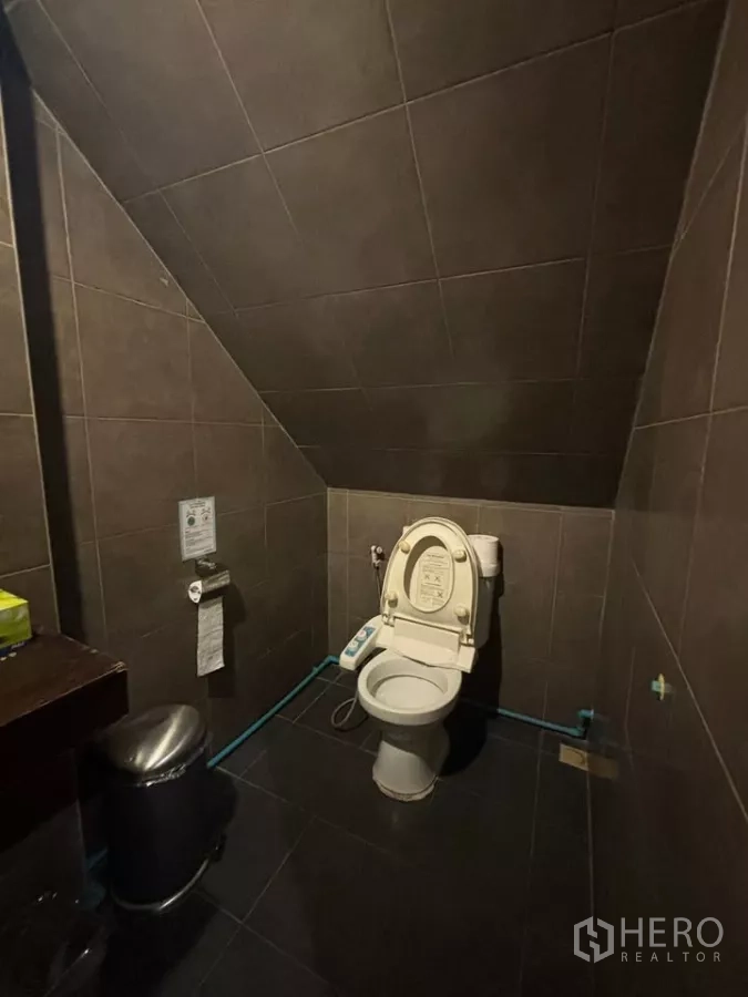 Shophouse for rent in Watthana, Bangkok - Under-stairs dark-tiled restroom with toilet and waste bin.