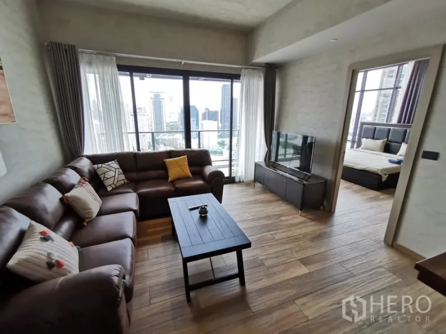 Condominium for rent in Watthana, Bangkok - Bright living room with leather sectional, TV console and floor-to-ceiling windows showcasing the Bangkok skyline, with a doorway to a bedroom.