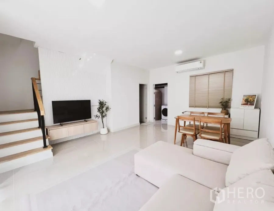 Townhouse for rent in Bang Phli, Samut Prakan - Bright open-plan living and dining area with sofa, wooden table, TV console and staircase in a modern Bangna townhouse.
