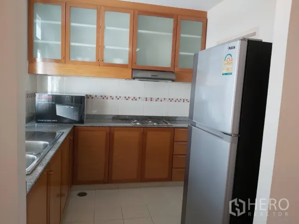 Apartment for rent in Khlong Toei, Bangkok - U-shaped kitchen with wood cabinets, granite countertops, stainless steel refrigerator, and double sink.
