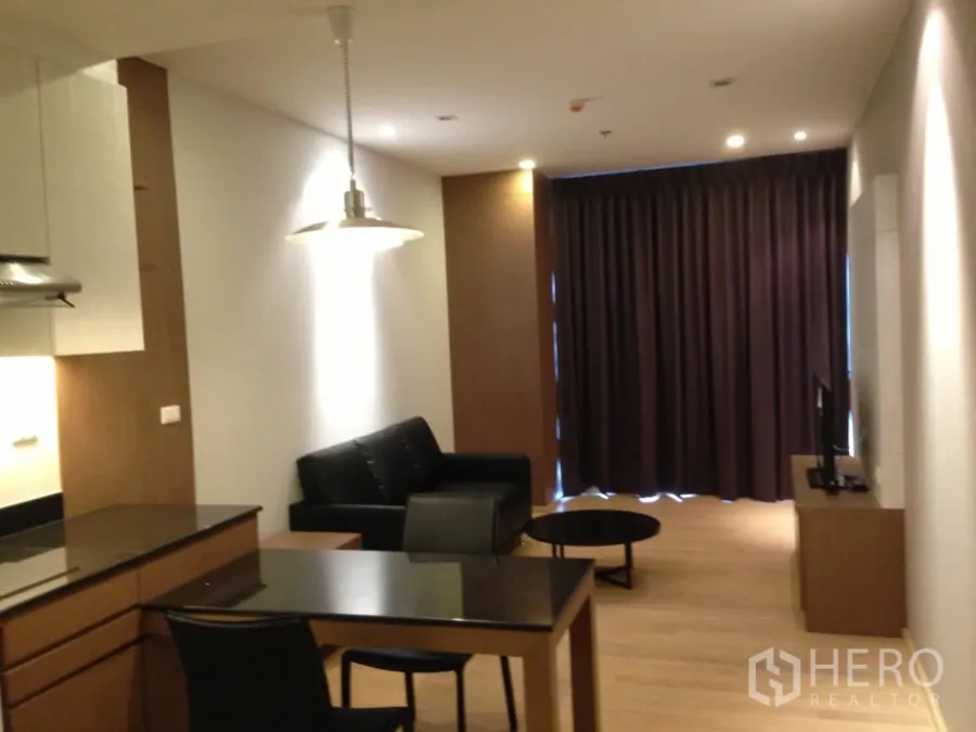 Condominium for rent in Bangkok - Open-plan living and dining area in a Khlong Toei condo with kitchenette, pendant light, black sofa and blackout curtains.