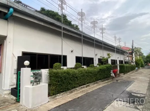 Factory for rent in Prawet, Bangkok - Side elevation of a low-rise factory building with hedge-lined frontage and multiple dark-framed windows.