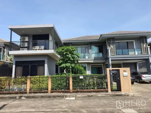 独立式住宅 出售在Suan Luang, Bangkok - 住宅正立面，设大面积二层露台、修剪整齐的绿篱与车道停车位。