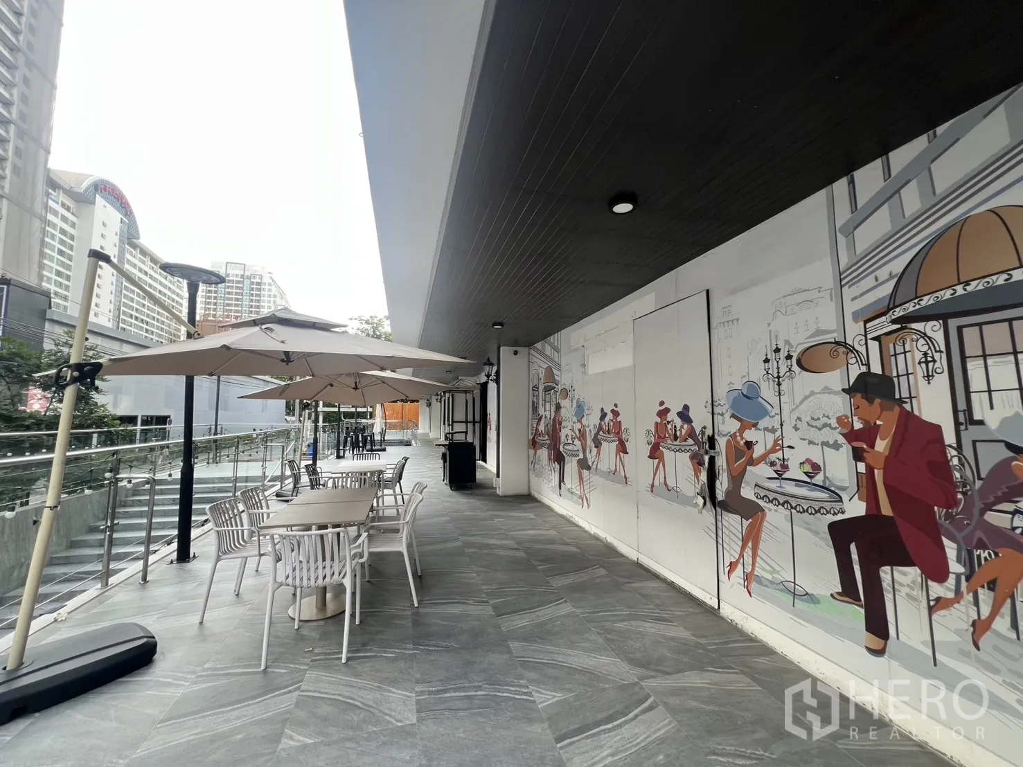 Retail Space for rent - Covered walkway with tables and parasols beside a colorful café mural, overlooking nearby city buildings.