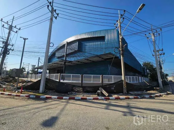 Factory for rent in Chon Buri - Corner view of a modern blue steel-clad factory with perimeter fencing and utility poles.
