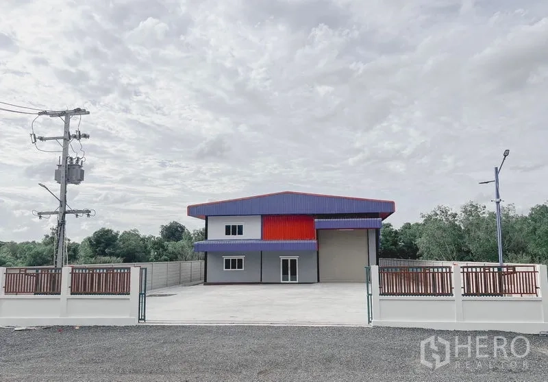 Warehouse for rent or sale in Khlong Luang, Pathum Thani - Front exterior of a modern gated warehouse with a wide concrete forecourt and two‑storey office, set amid greenery.