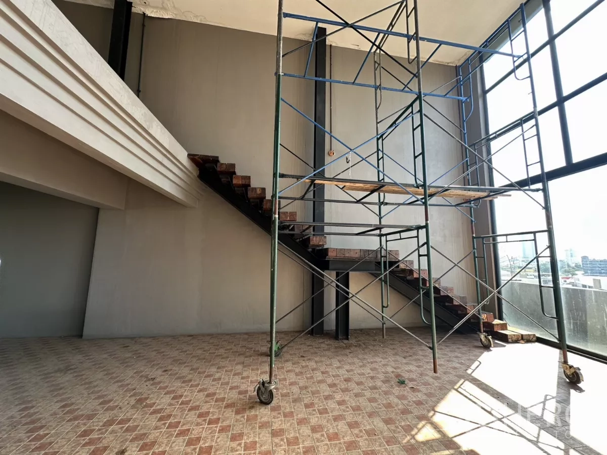 Shophouse for rent in Bang Na, Bangkok - High‑ceiling space with mezzanine stair and scaffolding beside full‑height windows.