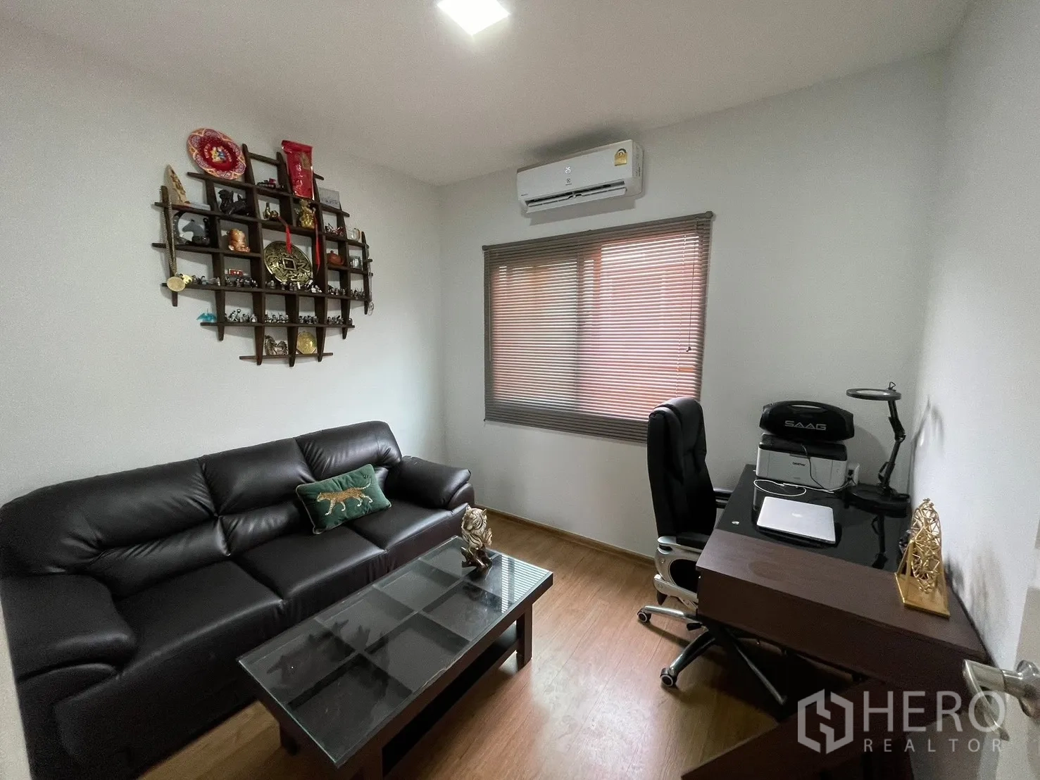 Townhouse for rent in Bang Phli, Samut Prakan - Compact living-room workspace with black leather sofa, glass coffee table, wall display shelf, and a desk beneath a window with blinds and air conditioning.