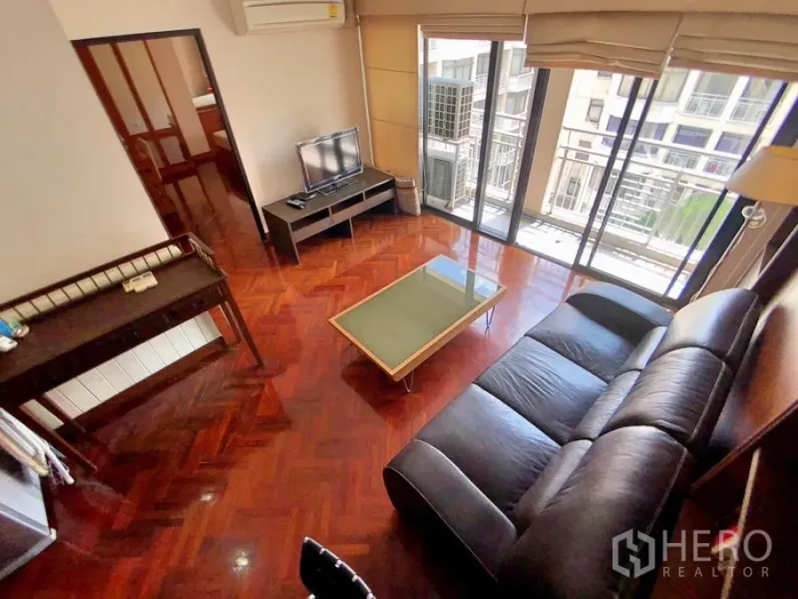 Condominium for rent in Bang Rak, Bangkok - Bright living room with black leather sofa, glass coffee table, TV console, and floor-to-ceiling windows opening to a balcony with city views.