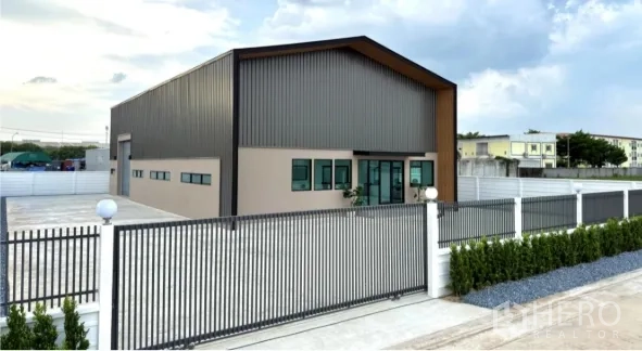 Warehouse for sale in Bang Bua Thong, Nonthaburi - Angled exterior view showing the gated entrance and wide concrete yard around the warehouse.