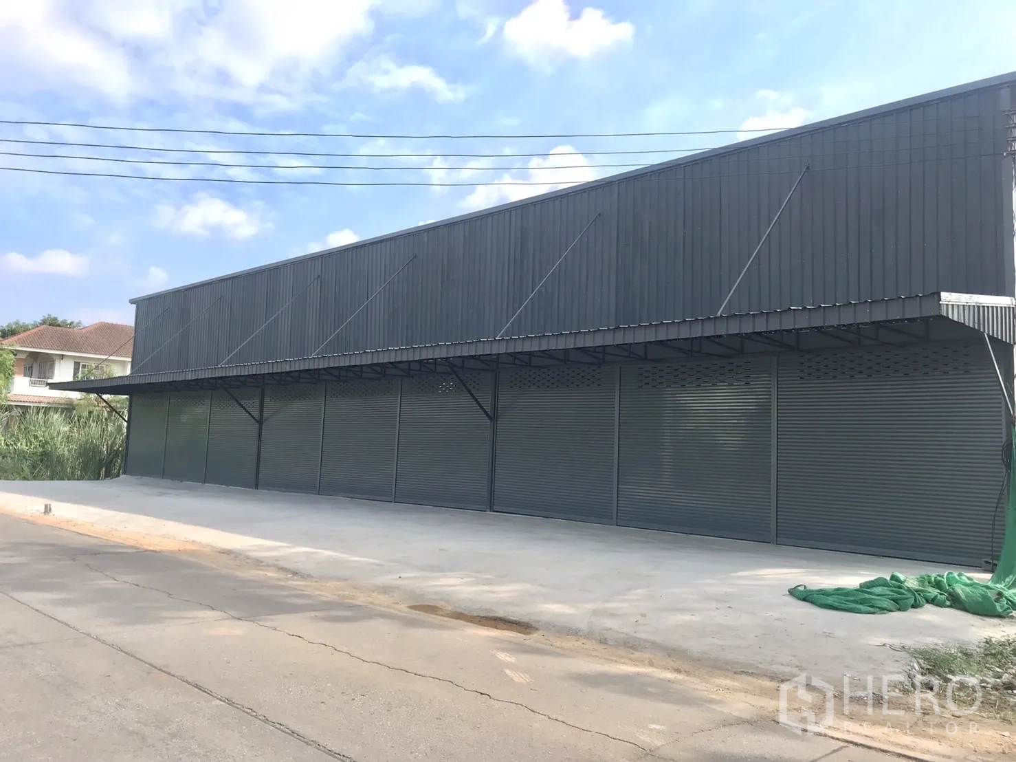 Warehouse for rent in Lam Luk Ka, Pathum Thani - Modern grey warehouse with multiple roller shutters and a wide concrete loading area facing the street.
