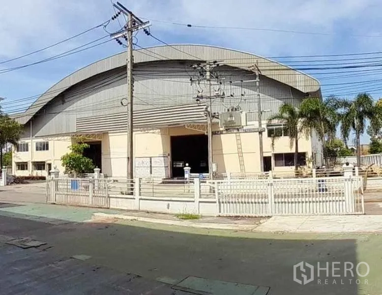 Factory for rent in Mueang Samut Prakan, Samut Prakan - Street view of a large curved‑roof factory with gated entrance, palm trees and overhead power lines.