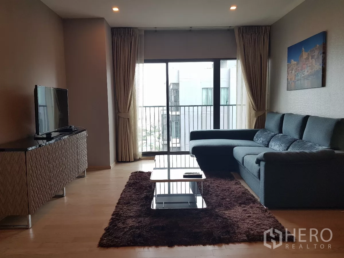 Condominium for rent in Khlong Toei, Bangkok - Bright living room with L-shaped sofa, TV console and sliding doors to a balcony with city view.