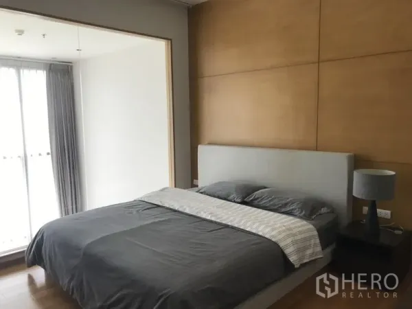 Condominium for rent in Khlong Toei, Bangkok - Bright bedroom with wood accent wall, upholstered bed, bedside table and curtained window.