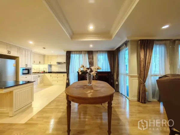 Condominium for rent in Pathum Wan, Bangkok - Dining table facing an open U-shaped kitchen with cream cabinets and large windows.
