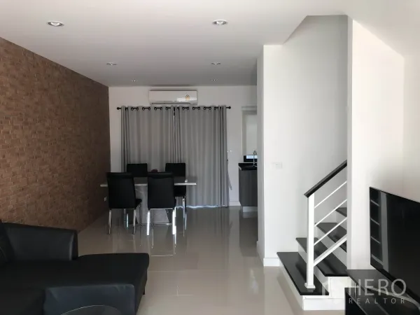 Townhouse for rent in Bang Kapi, Bangkok - Open-plan living and dining room with brick feature wall, glossy tiled floors, staircase and a compact kitchen beyond.