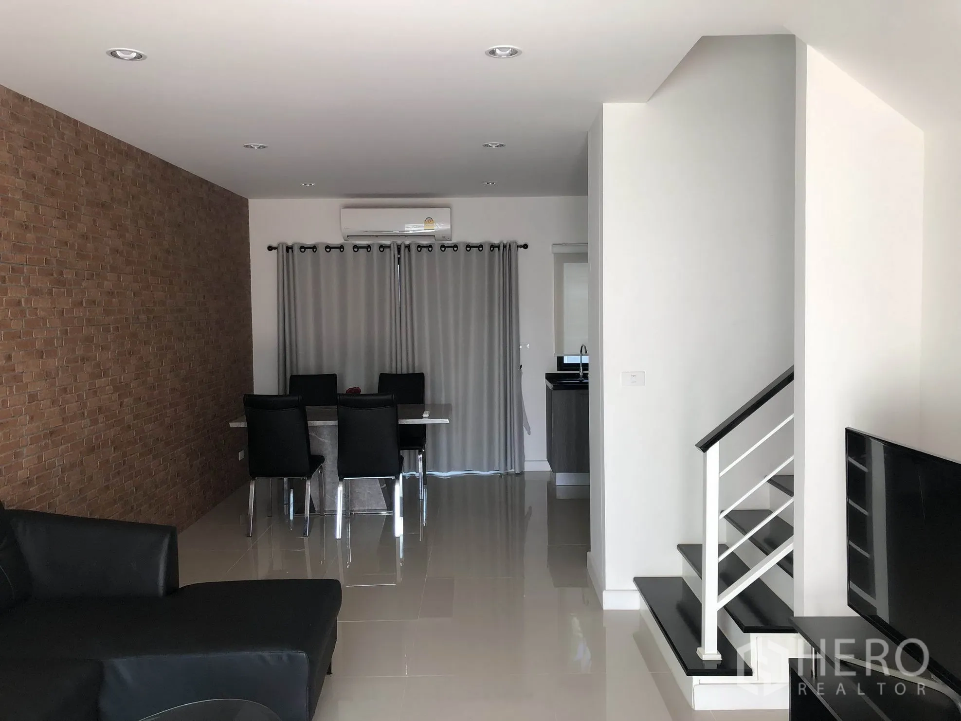 Townhouse for rent in Bang Kapi, Bangkok - Open-plan living and dining room with brick feature wall, glossy tiled floors, staircase and a compact kitchen beyond.