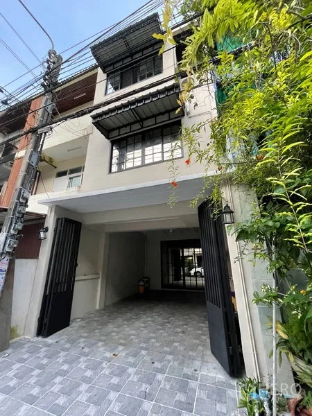 联排别墅 出租在Phra Khanong, Bangkok - 三层联排别墅外立面，带铁门车库与铺砖车道及绿植。