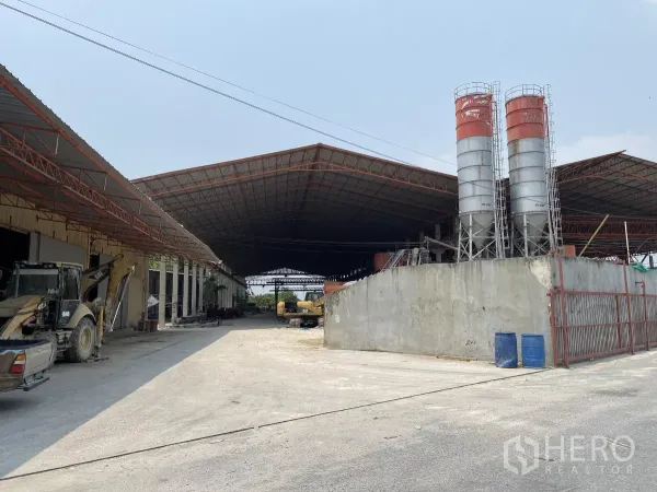 Factory for sale in Chon Buri - Expansive factory exterior in Nong Ri with wide steel-roofed bays, twin cement silos and heavy machinery at the entrance.