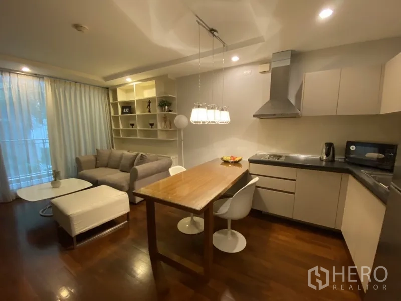 Condominium for rent or sale in Khlong Toei, Bangkok - Open-plan living room with built-in shelves, gray sofa, wood floors, and an L-shaped kitchen with stainless hood and pendant lights.