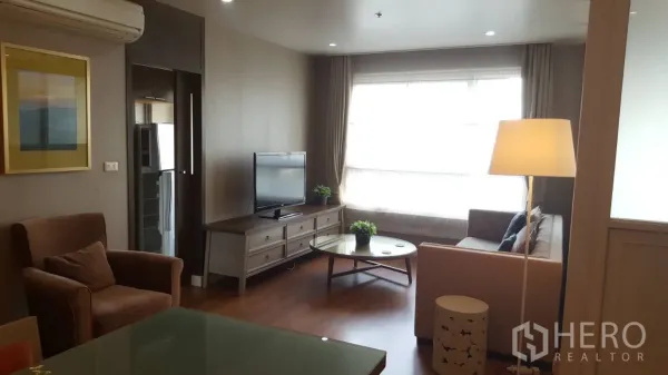 Condominium for rent in Khlong Toei, Bangkok - Bright condo living room with large window, sofa, TV console and round glass coffee table on wooden floors.