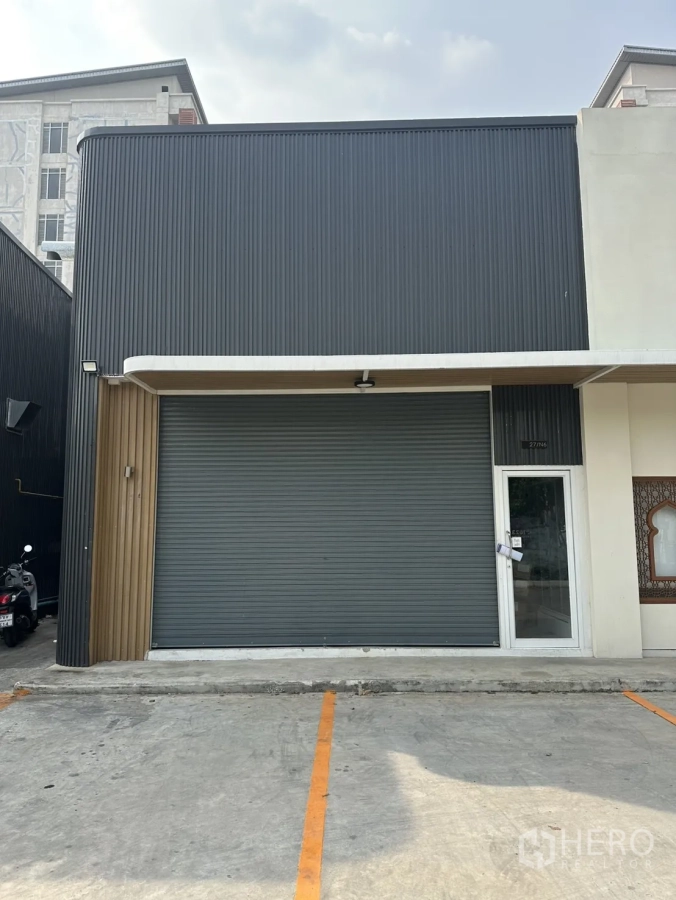 Warehouse for rent in Suan Luang, Bangkok - Modern warehouse exterior with corrugated metal cladding, wide roller shutter, glass entry door and marked parking bays.