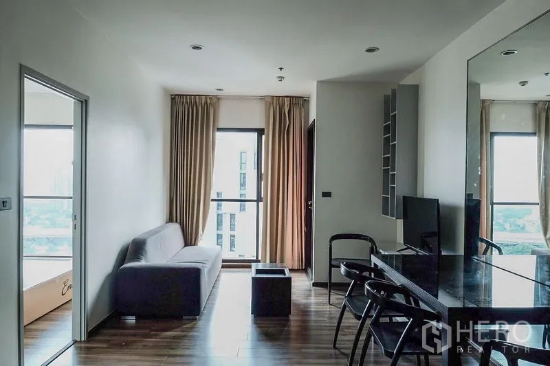 Condominium for rent in Khlong Toei, Bangkok - Bright living room with gray sofa, floor-to-ceiling window and mirror wall in a Bangkok condo.
