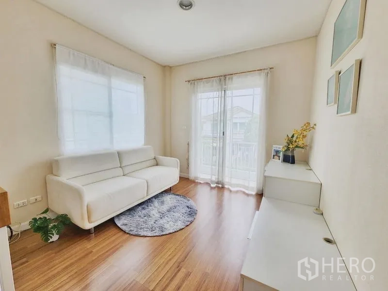 Detached House for rent in Prawet, Bangkok - Sunlit upstairs family room with white sofa, balcony doors and warm wood floors.