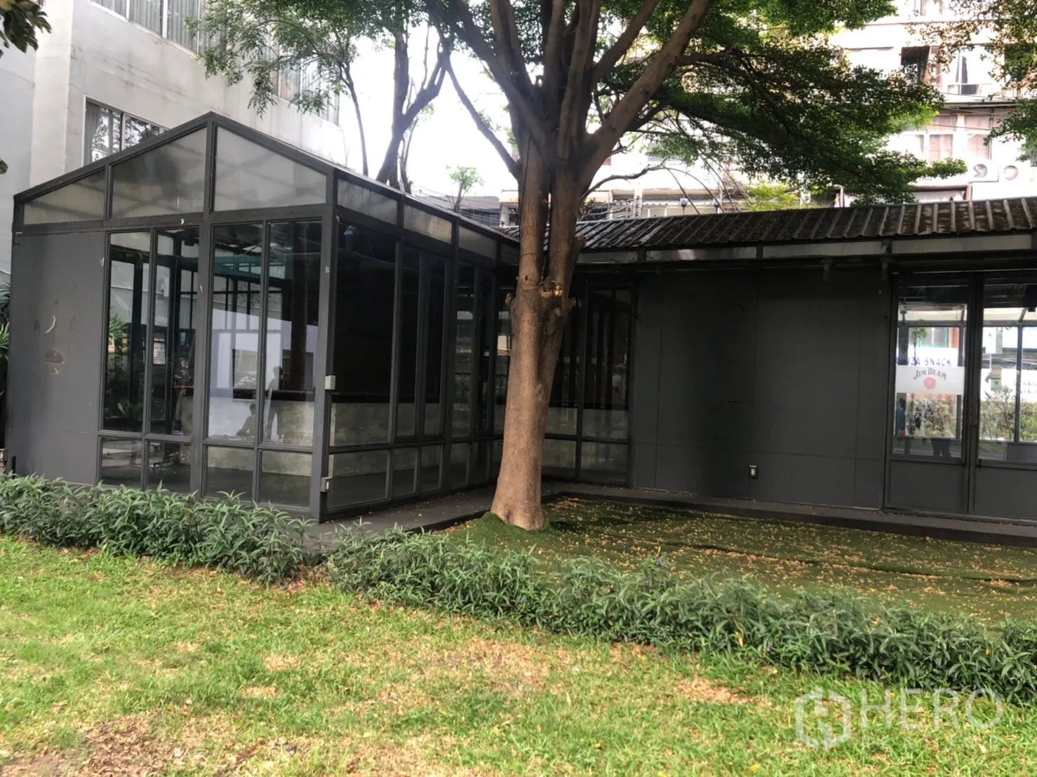 Retail Space for rent in Watthana, Bangkok - Exterior view of a glass pavilion and dark-clad building shaded by a tree with landscaped edging.