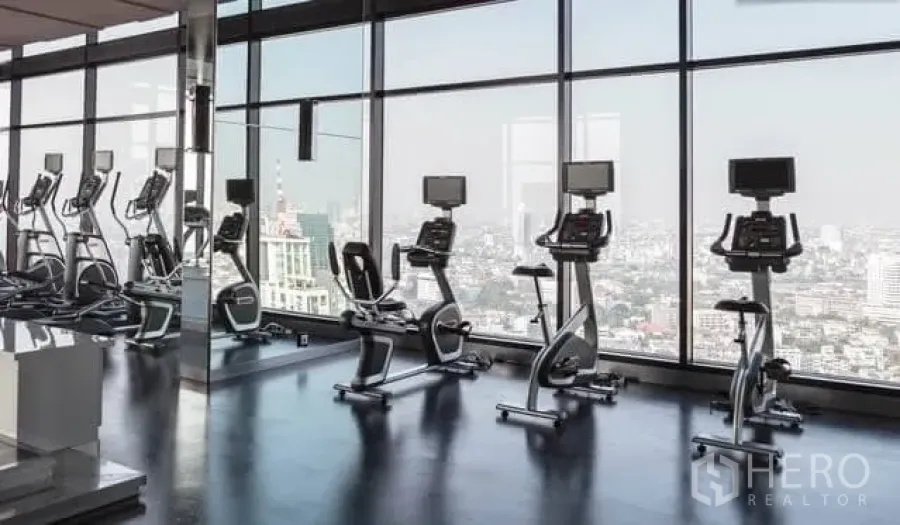 Condominium for rent in Khlong Toei, Bangkok - High-rise gym with cardio bikes facing floor-to-ceiling windows and Bangkok skyline.