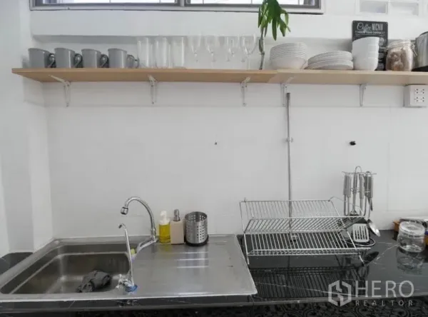 Townhouse for rent in Khlong Toei, Bangkok - Compact white kitchen featuring a stainless sink, open shelves with cups and glassware, and a countertop dish rack.
