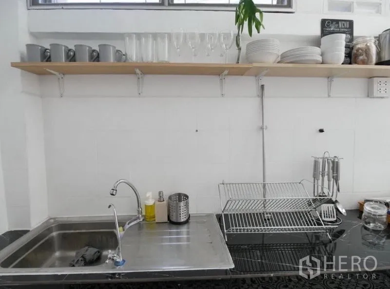 Townhouse for rent in Khlong Toei, Bangkok - Compact white kitchen featuring a stainless sink, open shelves with cups and glassware, and a countertop dish rack.
