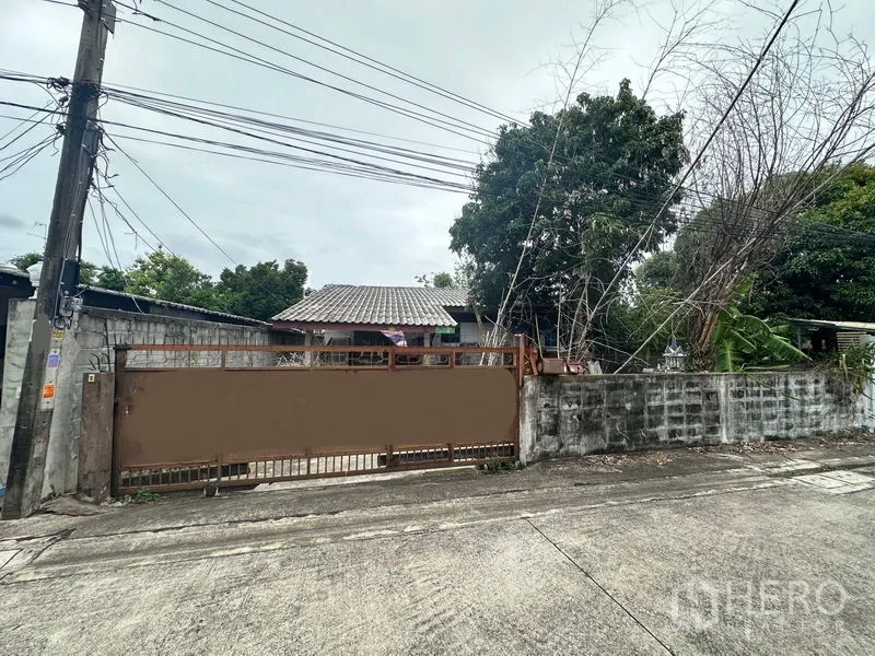 地 出售在Phra Khanong, Bangkok - 地块正面滑动金属大门与围墙，大树成荫，可见市政管线。