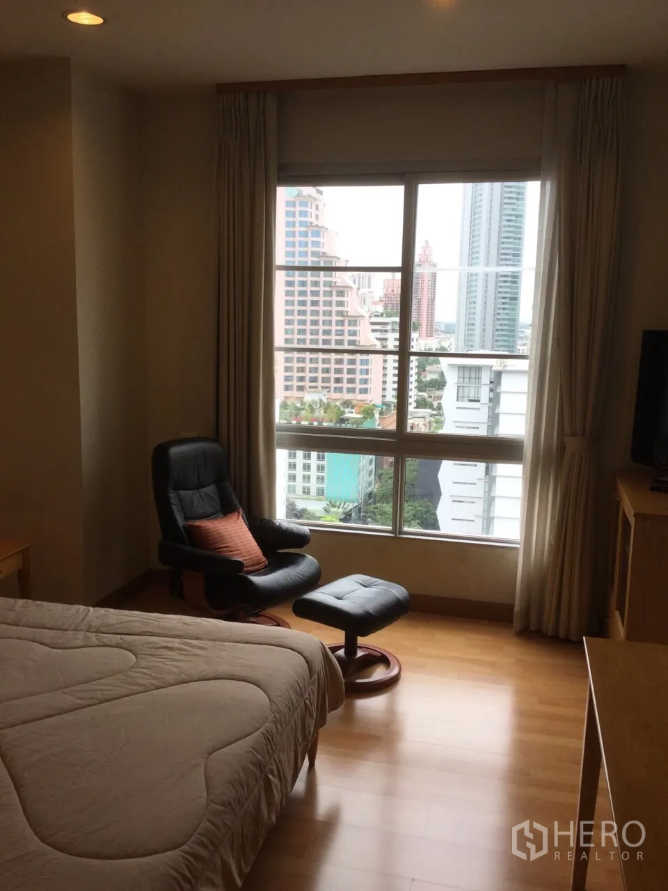 Condominium for rent in Khlong Toei, Bangkok - Bedroom with large window, city skyline view and leather lounge chair with ottoman.