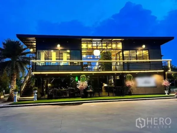 Detached House for sale in Khlong Luang, Pathum Thani - Modern two-storey glass building illuminated at night with balcony and outdoor seating.
