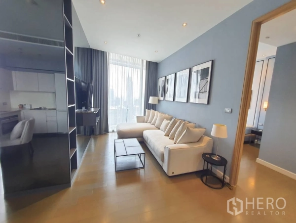 Condominium for rent in Pathum Wan, Bangkok - Open living space showing large sofa, TV wall, tall curtains and doorway leading to the bedroom.