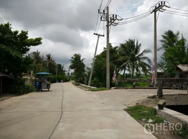 Factory for rent or sale in Krathum Baen, Samut Sakhon - Quiet local road with power lines and a small canal bridge amid lush greenery.