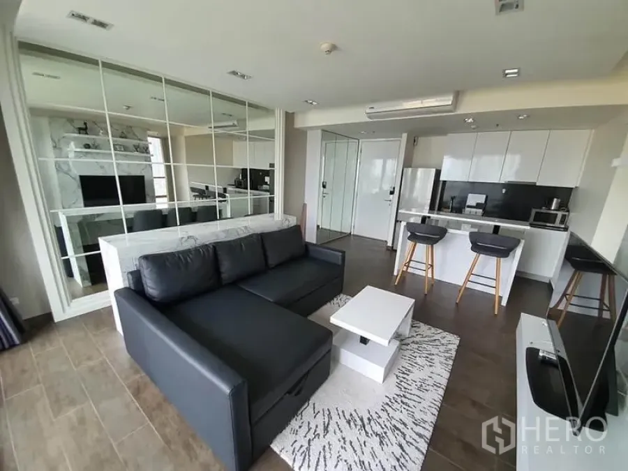 Condominium for rent or sale in Watthana, Bangkok - Open-plan living room with black leather sofa, mirrored feature wall, and a white island kitchen.