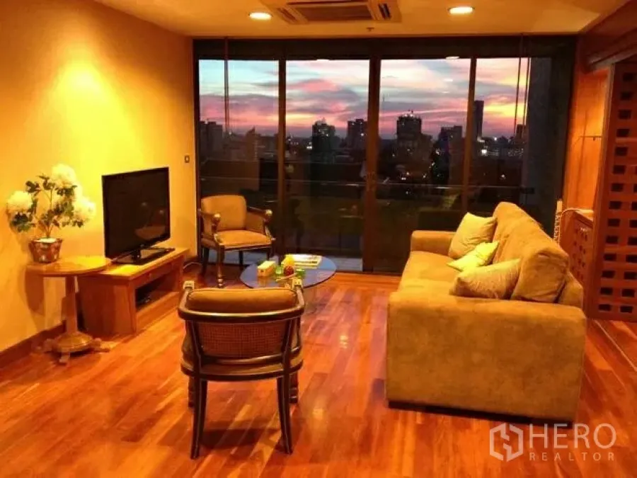 Condominium for sale in Pathum Wan, Bangkok - Warm-lit living room with sofa set, TV, hardwood floors, and wide windows framing the skyline at sunset.