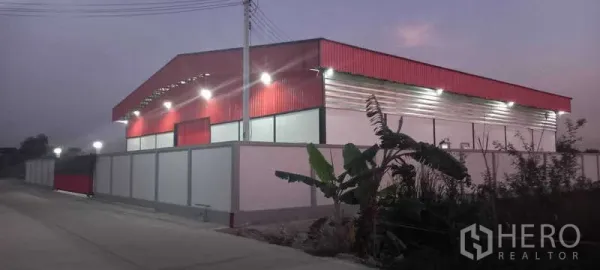 Warehouse for rent or sale in Khlong Luang, Pathum Thani - Evening side view of a red metal-clad warehouse behind a perimeter wall with street lighting along a quiet road.