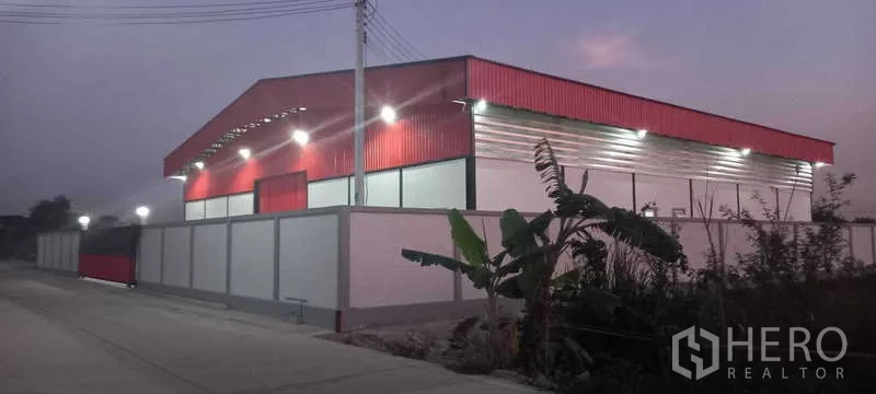 Warehouse for rent or sale in Khlong Luang, Pathum Thani - Evening side view of a red metal-clad warehouse behind a perimeter wall with street lighting along a quiet road.
