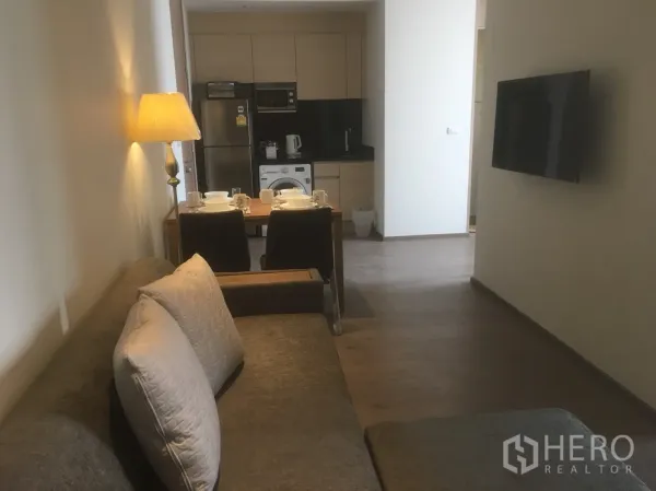 Condominium for rent or sale in Khlong Toei, Bangkok - Open-plan living and dining area with sofa, wall-mounted TV and kitchen beyond.