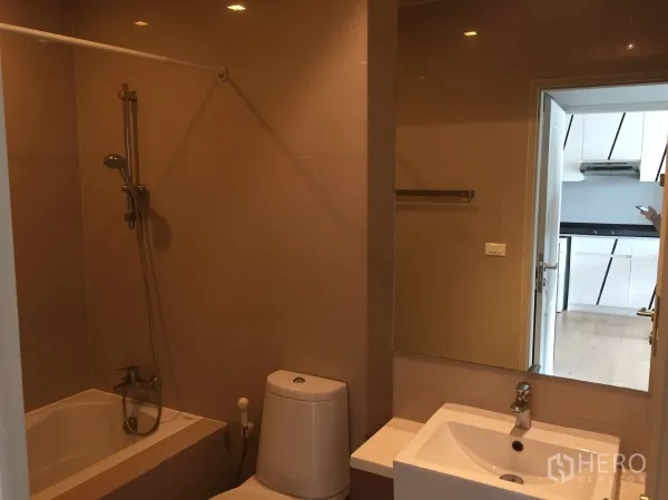 Condominium for rent in Bangkok - Modern bathroom with bathtub and shower combo, large mirror, toilet and vanity set against beige tiles.